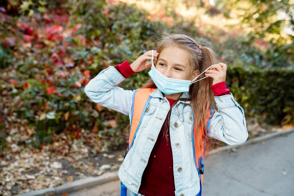Back to school. A 7-year-old girl wearing a face mask goes to school along an alley in the park in autumn - Foto, imagen