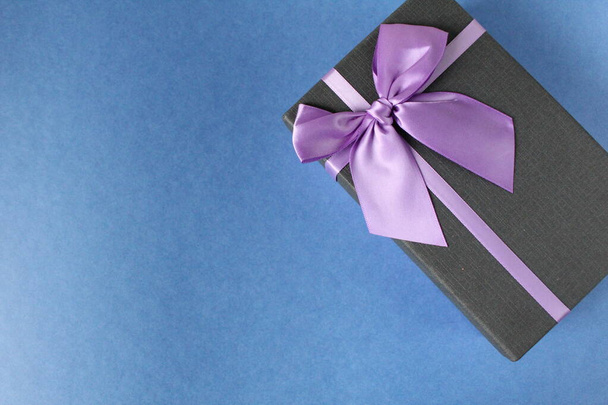 dárek v černé krabici a fialové šeříkové stuhy a luk na dovolenou Nový rok Vánoce Den matek narozeniny firemní večírek na modrém pozadí s kopírovacím prostorem - Fotografie, Obrázek