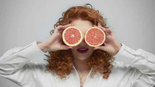 Девушка покрывает лицо половинками грейпфрута. Леди позирует в студии с грейпфрутом - Фото, изображение