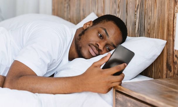 Розроблений африканський хлопець дивлячись на мобільний телефон лежить в ліжку
 - Фото, зображення