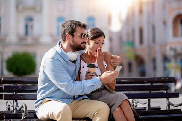 Jeune couple assis dans la ville, boire du café à emporter, regarder son téléphone intelligent et profiter de la journée ensoleillée.  - Photo, image