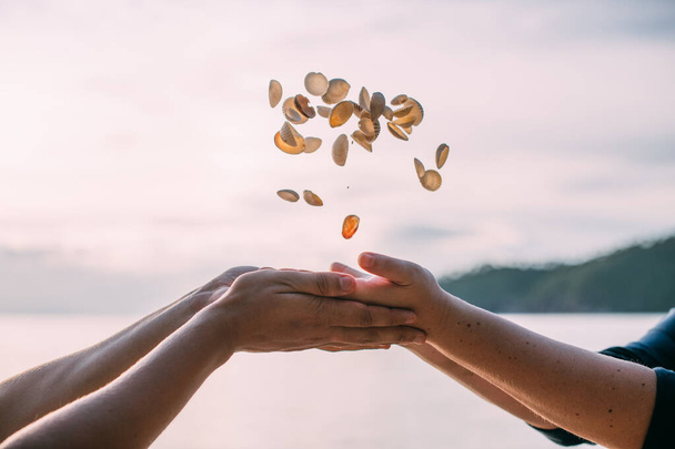 Male and female hands toss a full handful of shells at sunset. Close-up. Small white shells fly over the palms of a man and a woman against the background of the sea - Photo, Image