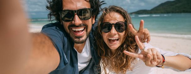Rakastavaiset ottavat selfien trooppisella rannalla. Mies ja tyttö hymyilevät iloisesti puhelimen kameralle meren taustalla. Vastanaineet ja trooppinen laguuni vihreillä vuorilla ja pilvisellä taivaalla
. - Valokuva, kuva