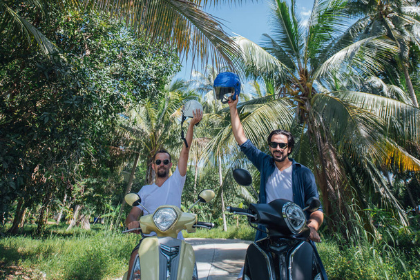 Двое мужчин на мотоциклах со шлемами в пальмах на тропическом острове. Ребята туристы на велосипедах по дороге в пальмовой роще
 - Фото, изображение