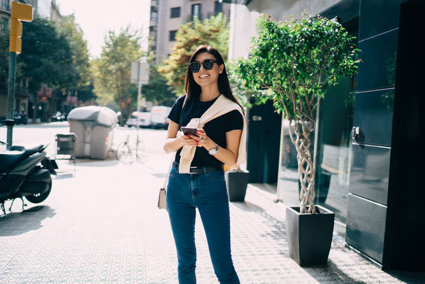 Half length portrait of smiling asian woman in casual wear and sunglasses using modern smartphone for chatting, positive female 20s holding mobile phone sending text messages and mails on street - Fotó, kép