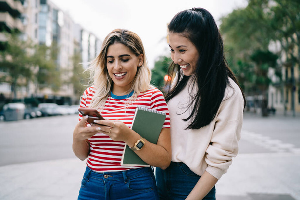 Joyful hipster girls reading funny publication during social networking via modern cellular technology enjoying together blogging in city, smiling female tourists tracking mobile gps during travelling - Фото, зображення