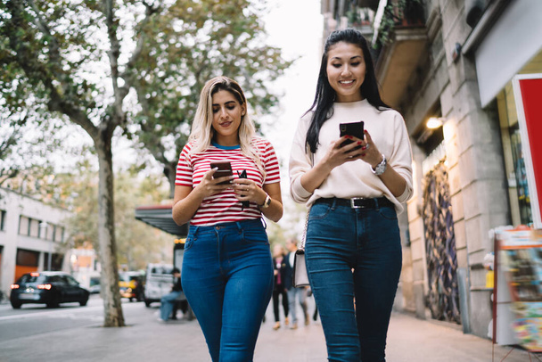 Multiracial female tourists with cellphone gadgets in hands walking at urban setting and ignoring live communication during together travel vacations, addicted hipster girls chatting in networks - Foto, Bild