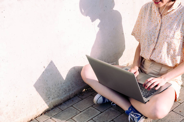 felismerhetetlen fiatal nő fülhallgatóval dolgozik egy számítógéppel ül egy szürke fal mellett, technológia koncepció és blogolás életmód, szövegmásolási hely - Fotó, kép