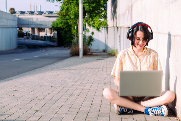 front view of a young woman with headphones working with a computer sitting next to a gray wall, technology concept and urban lifestyle, text copy space - Fotó, kép