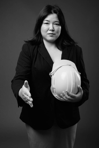 Красивая толстая азиатская бизнесвумен держит каску на сером фоне - Фото, изображение