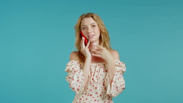 Pretty young woman with blonde hair speaks, smiling on mobile phone. Beautiful girl holding and using smart phone. Cute floral outfit. Blue studio background. - Video, Çekim