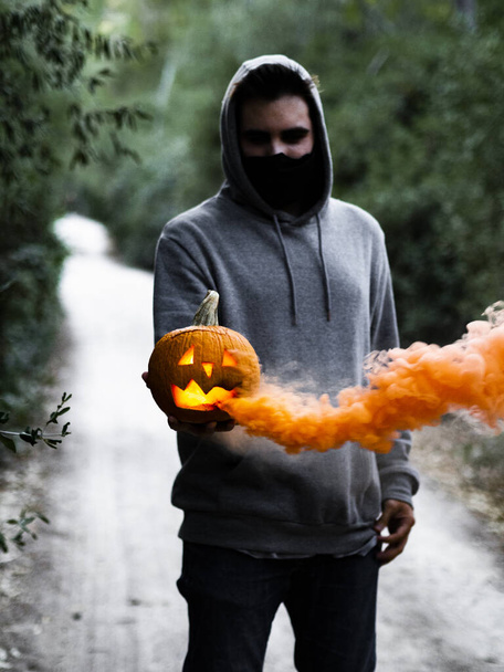 A spooky shot of a European guy holding a smoking Halloween pumpkin while wearing a facemask - 写真・画像