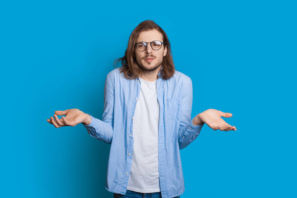 Confuso uomo caucasico con barba e capelli lunghi posa indeciso mentre gesticola con le palme su un muro di studio blu - Foto, immagini