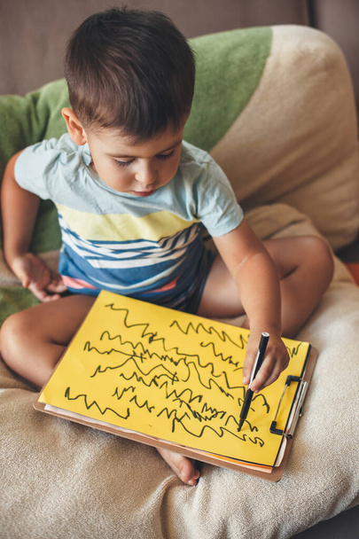 Lindo chico caucásico está dibujando algo en un papel amarillo con un marcador mientras está sentado en casa en la cama - Foto, imagen