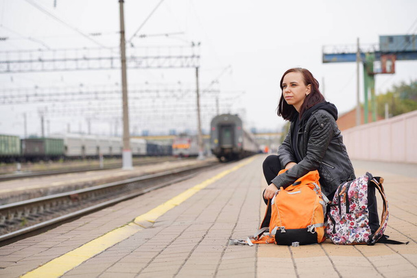 Young woman squats on platform, waiting for train. Female passenger with backpacks sitting on railroad platform in waiting for train ride - Foto, afbeelding