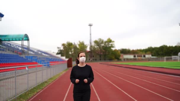 Active woman at stadium. Young fit female in sportswear and protective mask for coronavirus prevention running on red track during outdoor workout at stadium. - Video