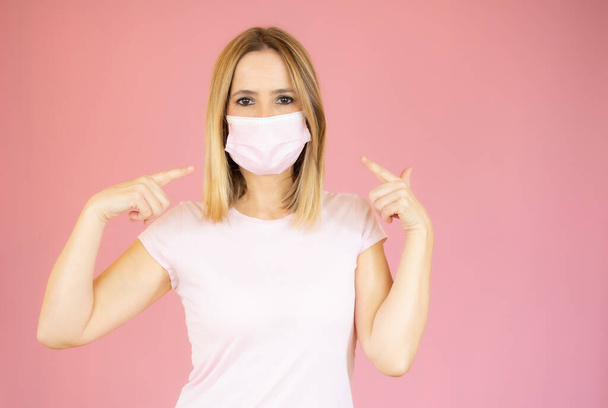 Blanke vrouw draagt een gezichtsmasker en wijst ernaar in een studio met een roze achtergrond - Foto, afbeelding