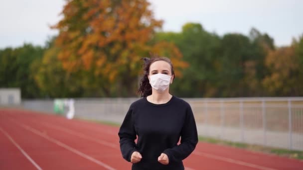 Active woman at stadium. Young fit female in sportswear and protective mask for coronavirus prevention running on red track during outdoor workout at stadium. - Video, Çekim