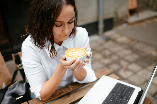 mulher segurando xícara de café nas mãos com laptop na mesa no café ao ar livre - Foto, Imagem