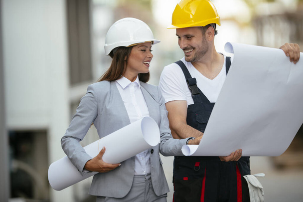 uśmiechnięty budowniczy mężczyzna i kobieta architekt w kaskach trzymających papierowe plany - Zdjęcie, obraz