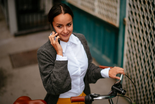 hymyilevä nuori nainen puhuu puhelimessa seistessään polkupyörän vieressä - Valokuva, kuva