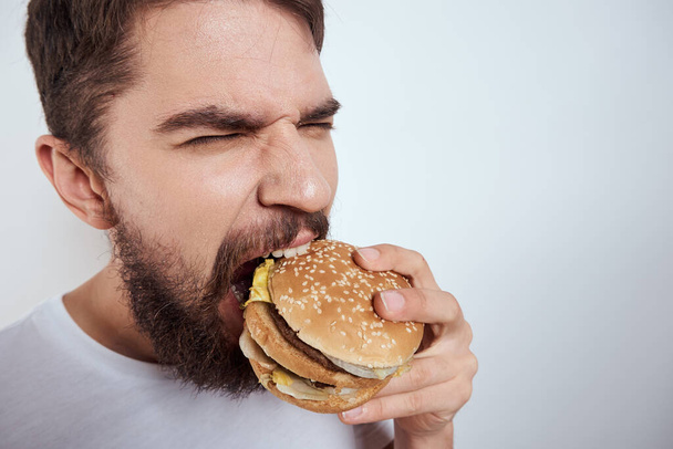Egy férfi hamburgert eszik könnyű háttérrel fehér pólóban kivágott szemmel közelről éhség gyorsétterem - Fotó, kép