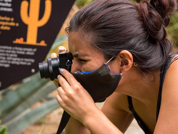 Junge Frau mit braunem Haar und schwarzer Gesichtsmaske fotografiert mit schwarzer Kompaktkamera - Foto, Bild
