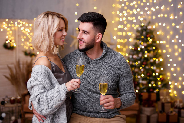 Концепция Рождества, Нового года и любви - портрет красивой пары, пьющей шампанское в украшенной гостиной с елкой и огнями - Фото, изображение