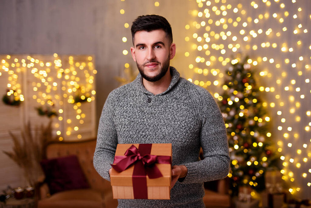 Weihnachts- und Neujahrskonzept - Porträt eines gutaussehenden bärtigen Mannes mit Geschenkbox im geschmückten Wohnzimmer mit Weihnachtsbaum und Lichtern - Foto, Bild