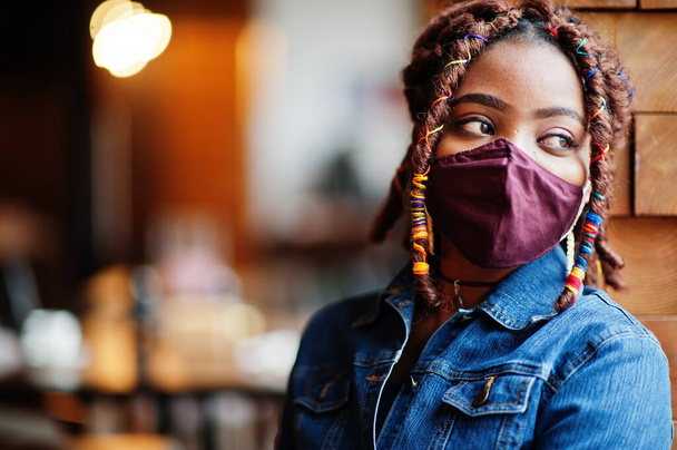 Stylish african american woman with dreadlocks afro hair, wear jeans jacket and face protect  mask at restaurant. New normal life after coronavirus epidemic. - Фото, изображение