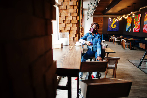 Stylish african american woman with dreadlocks afro hair, wear jeans jacket and face protect  mask at restaurant, hold cellphone. New normal life after coronavirus epidemic. - Fotografie, Obrázek