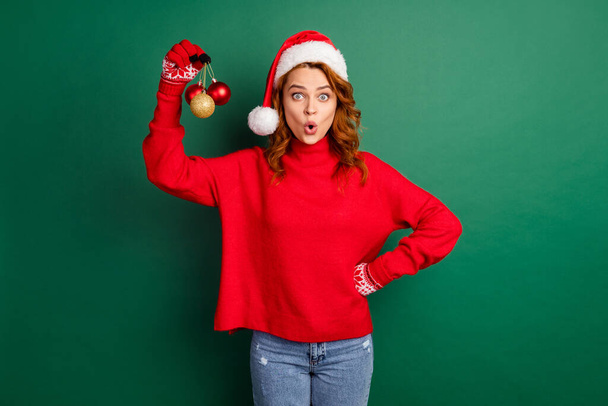 Photo of cute young lady hold demonstrate three baubles wear red sweater x-mas cap jeans isolated green color background - Фото, зображення