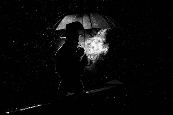 Silhouette eines Mannes mit Hut unter einem Regenschirm, der nachts im Regen in der Stadt eine Zigarette raucht - Foto, Bild