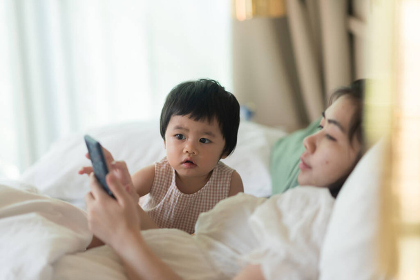 Güzel bebek annesiyle cep telefonuna bakıyor ve yatakta oturuyor. - Fotoğraf, Görsel