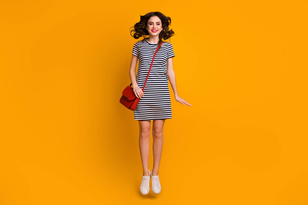 Full length photo of pretty curly millennial lady jumping high up sunny weather day for shopping wear white casual striped short dress clutch footwear isolated yellow color background - Foto, immagini