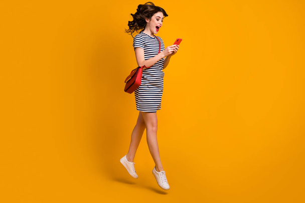 Full size profile photo of funny lady jump high read telephone sale season opening advert open mouth wear casual striped short dress clutch shoes isolated yellow color background - 写真・画像