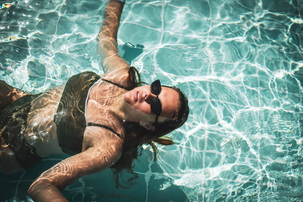 Модный портрет изящной милой девушки в стильных солнцезащитных очках, позирующих в бассейне во время отдыха на роскошном курорте. Весело плюс размер девочка-подросток наслаждается. счастливая, позитивная эмоция, летний стиль. - Фото, изображение