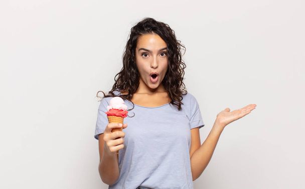 young hispanic woman looking surprised and shocked, with jaw dropped holding an object with an open hand on the side - Фото, изображение