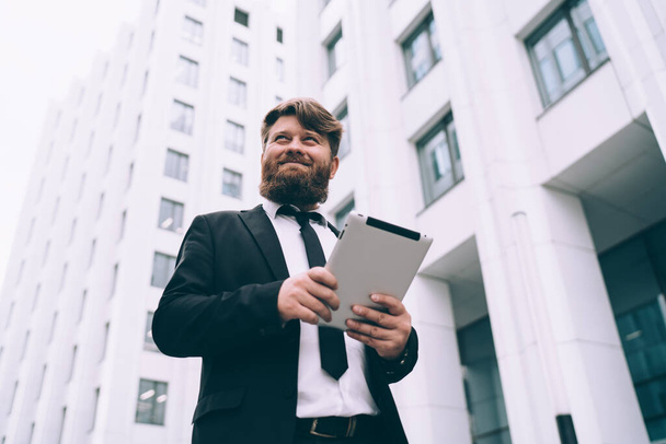 Low angle of bearded male with brown hair in black suit with tie walking with tablet on street with white buildings behind - Photo, Image