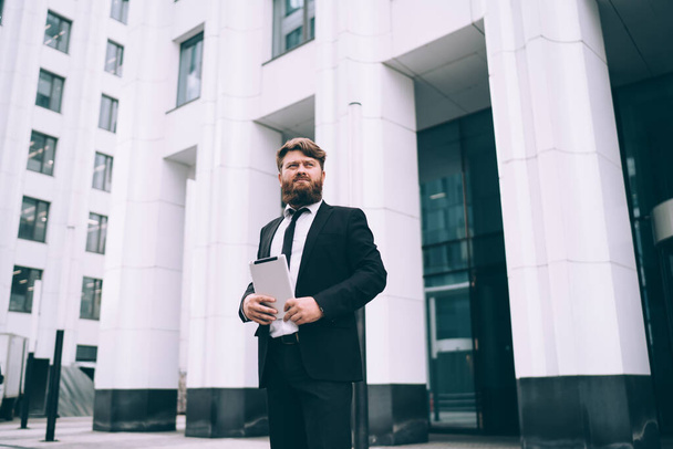 Confident bearded man with brown hair in black suit and tie standing on street near office building and holding tablet - Foto, Imagen