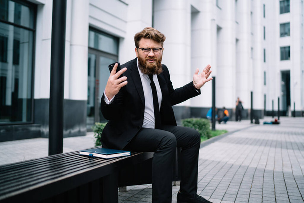 Young businessman in formal suit with beard and glasses explaining things and actively gesticulating while sitting in yard of office building - Foto, imagen