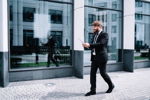 Side view of full length pensive bearded businessman in elegant clothes and tie checking documents while walking along pavement near modern office - Photo, Image