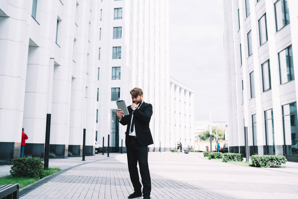 Full body of male leader standing on sidewalk near skyscraper with tablet looking information about financial news during online work - Foto, Imagen