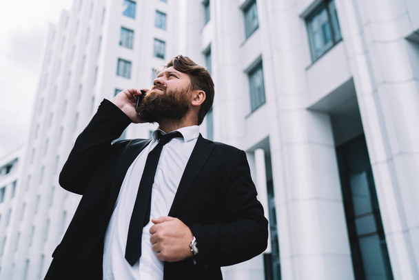 Serious entrepreneur in formal wear with beard discussing work issues on phone and looking up while standing in yard of office building - Foto, imagen