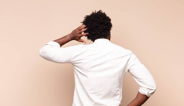 young black afro man feeling clueless and confused, thinking a solution, with hand on hip and other on head, rear view - 写真・画像
