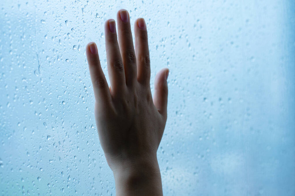 Γυναικείο χέρι στο παράθυρο κατά τη διάρκεια της βροχής. Γυαλί σε σταγόνες νερού - Φωτογραφία, εικόνα