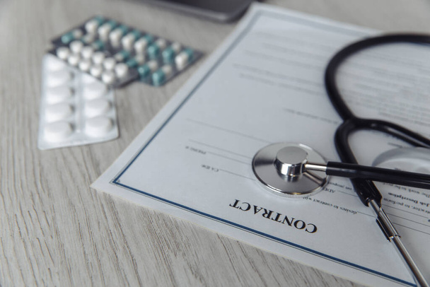Modern doctors office desk. Stethoscope and healthcare contract on wooden table close-up - Photo, Image