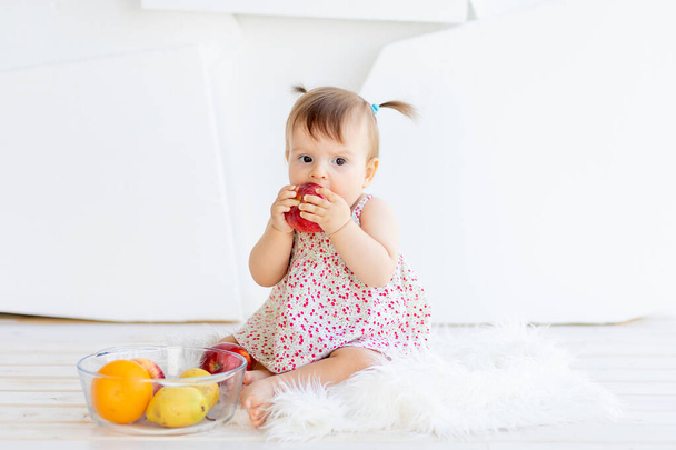 ein kleines Mädchen sitzt in einem hellen Raum mit einem Teller Obst und isst einen Apfel - Foto, Bild
