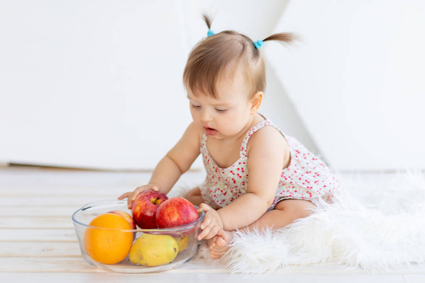 Parlak bir odada elinde meyve tabağıyla oturan küçük bir kız. - Fotoğraf, Görsel