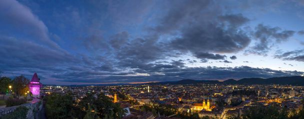 City lights of Graz and the famous clock tower on Schlossberg hill, Graz, Styria region, Austria, after sunset. Dramatic sky, panoramic view - Photo, Image